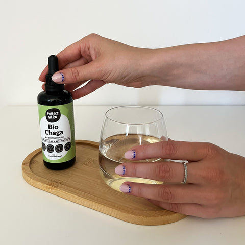 Hand holding a bottle labeled 'Bio Chaga' above a glass of liquid on a wooden tray.